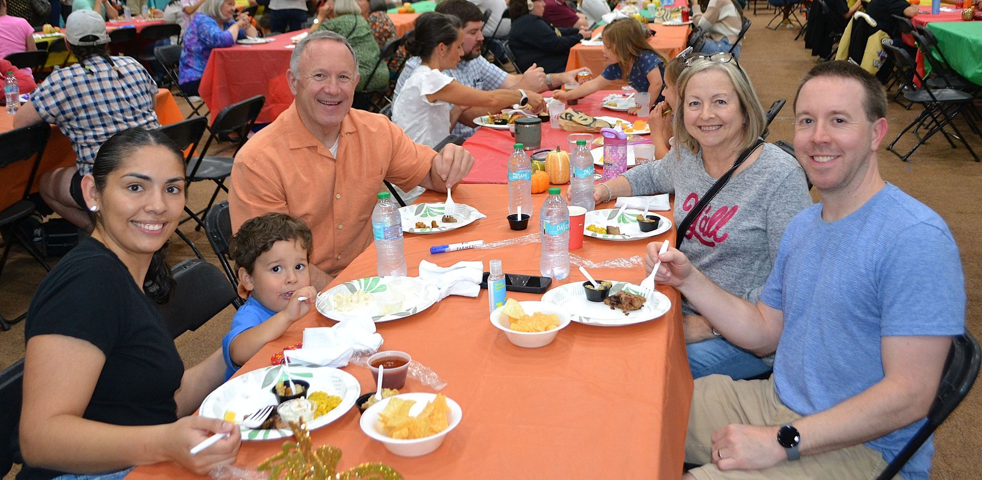 People gathered around a long table with orange tablecloths, sharing a meal and smiling at the camera.