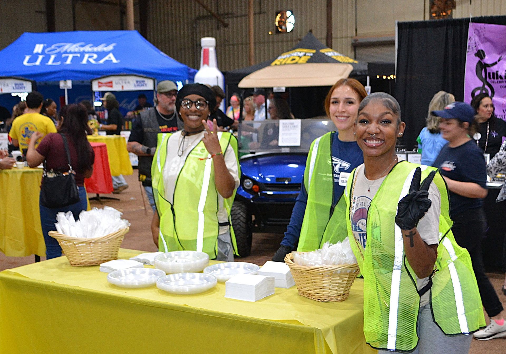Three volunteers in neon vests smiling behind a yellow table at an indoor event booth