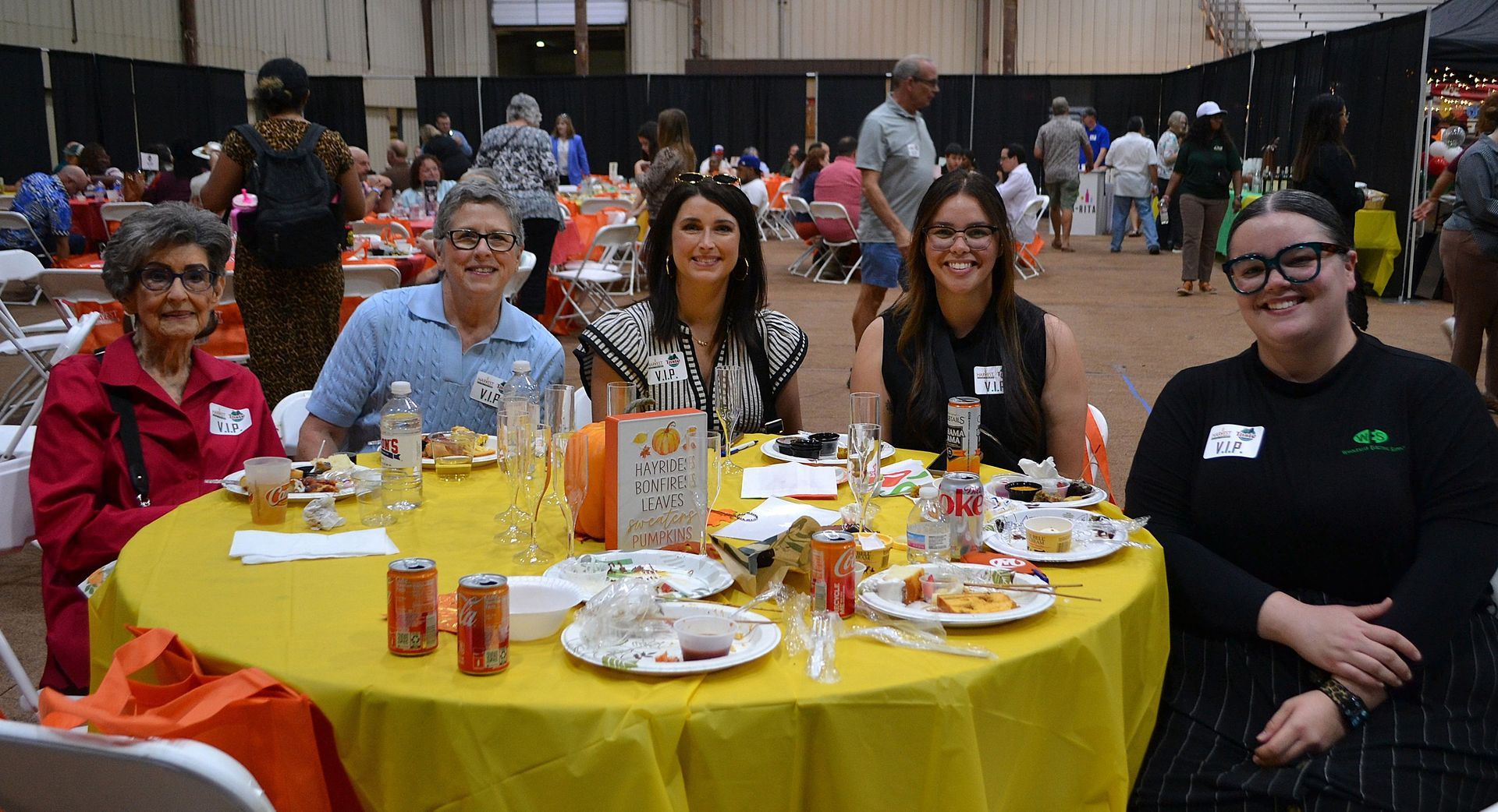 Five people seated at a yellow table with food and drinks in a busy indoor event hall