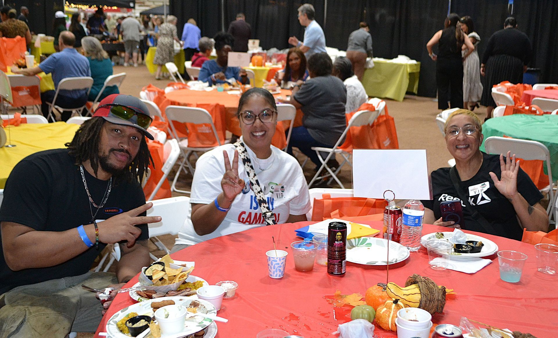 Three people smiling and waving at a table with food and drinks in a busy indoor event hall