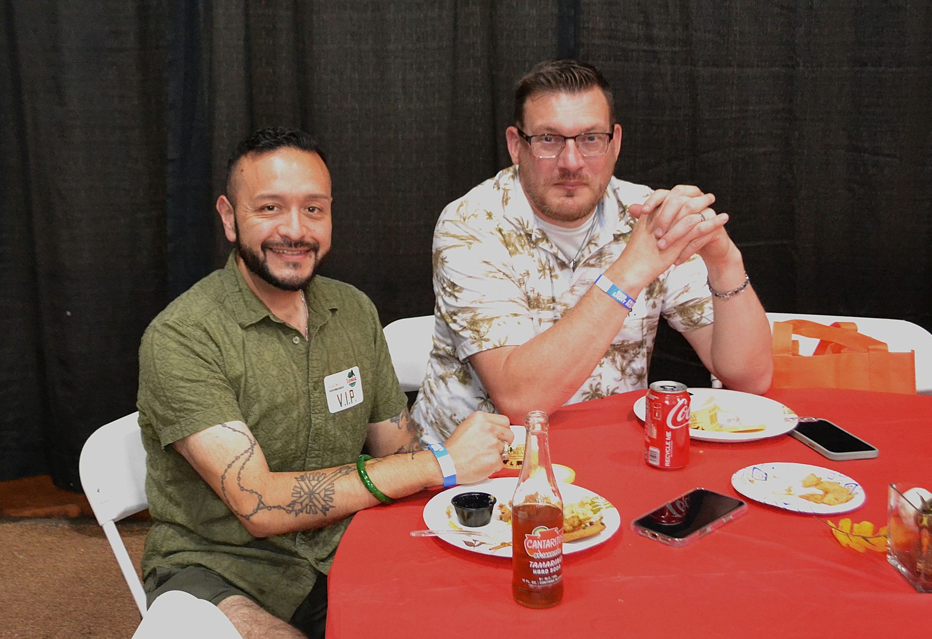 Two men seated at a red table with plates of food and drinks at an indoor event