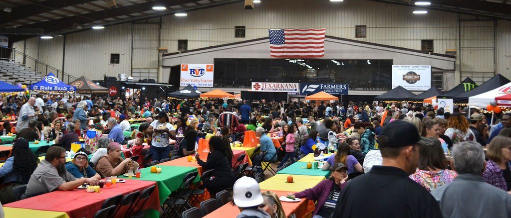 Crowded indoor event hall with rows of tables, people seated and walking, and a large American flag at the front