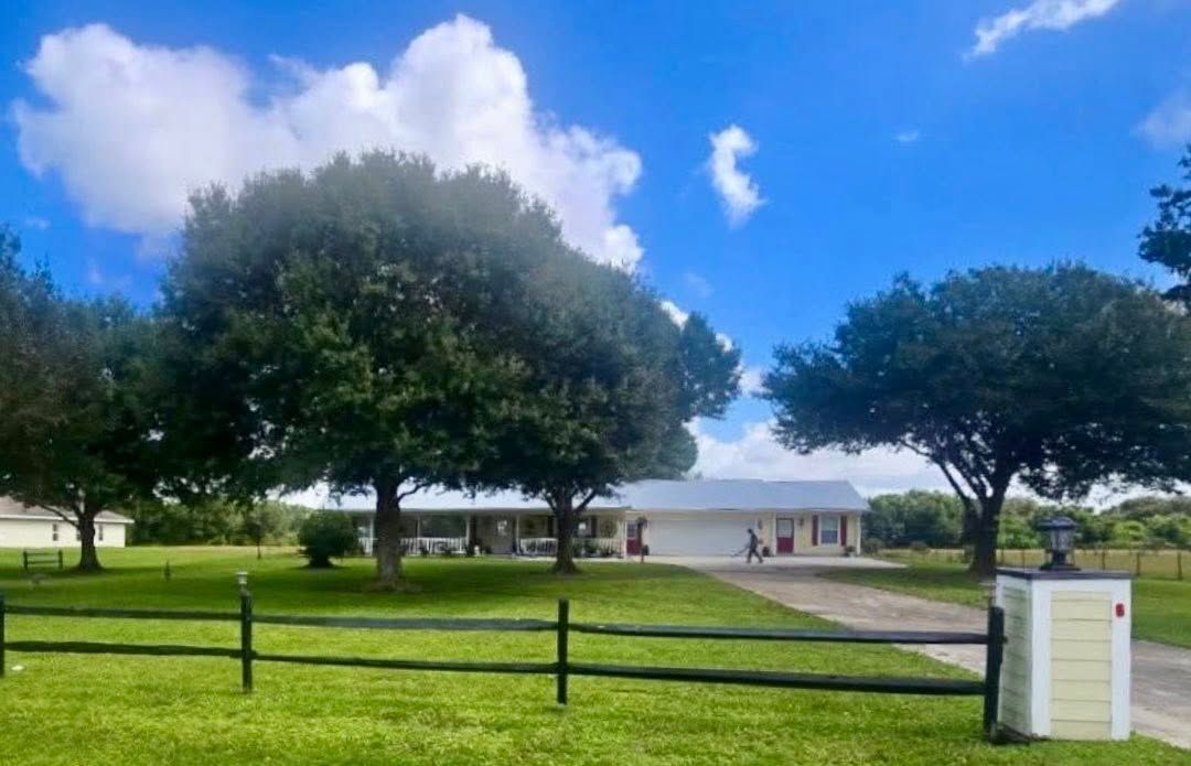 Green park with large trees, a white building, black fence, and bright blue sky.