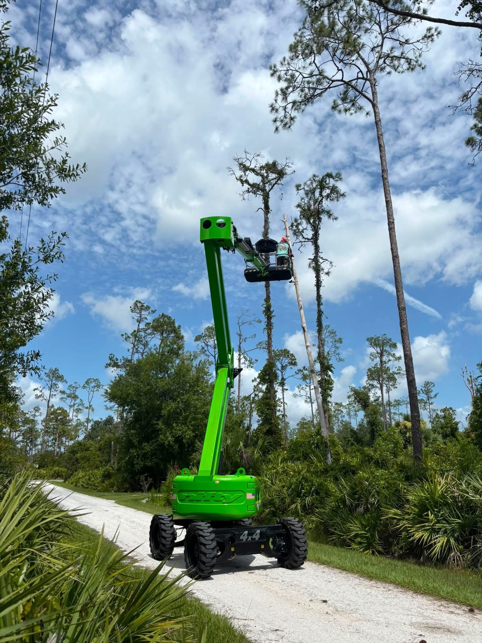 Green boom lift on a gravel path in a wooded area under a cloudy sky