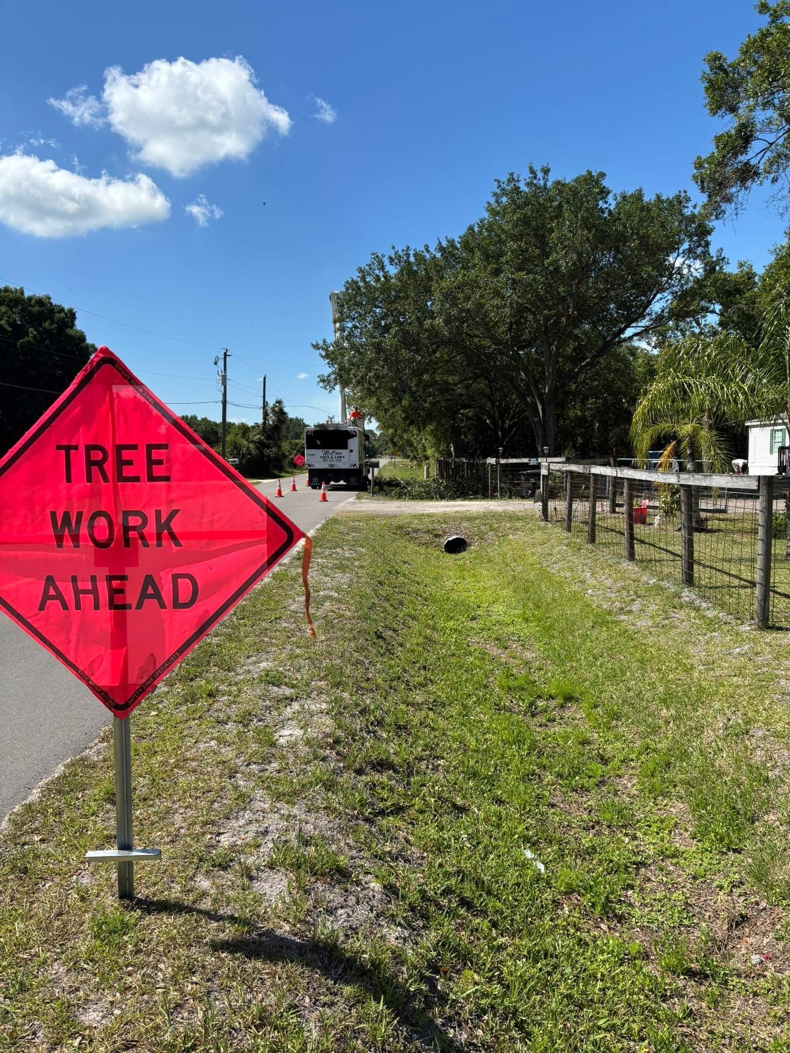 Red tree work ahead sign beside a roadside with trucks, fence, and grass under a blue sky