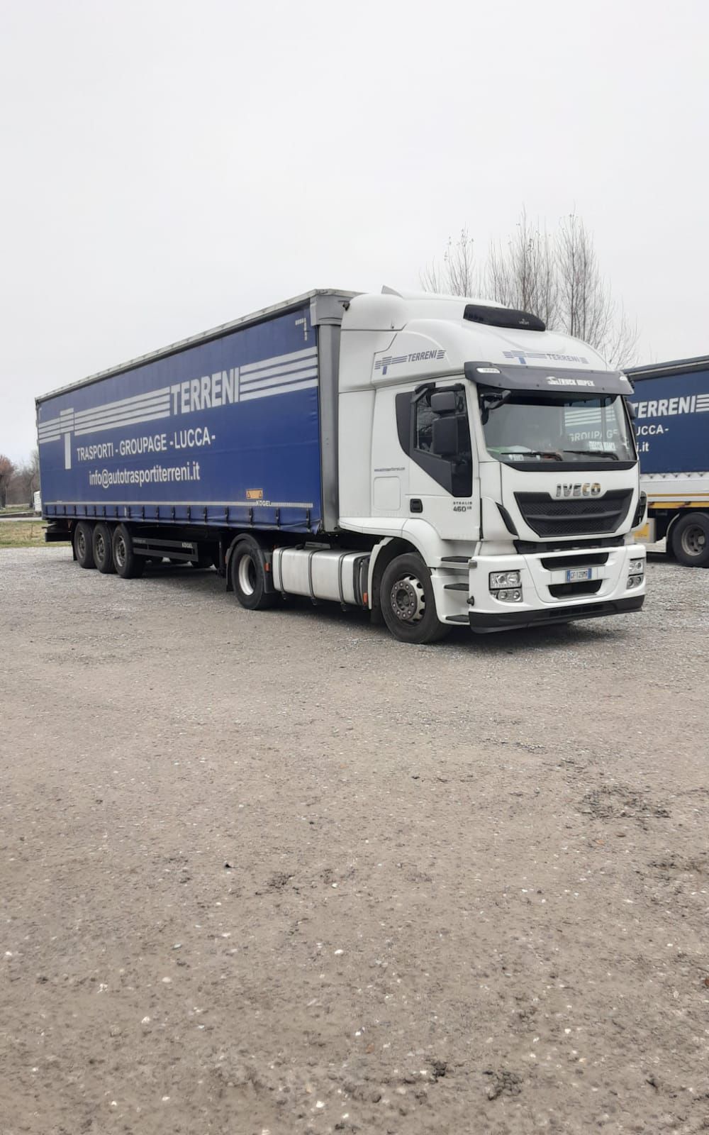 Un camion Iveco bianco con rimorchio blu parcheggiato su un piazzale ghiaioso sotto un cielo nuvoloso.