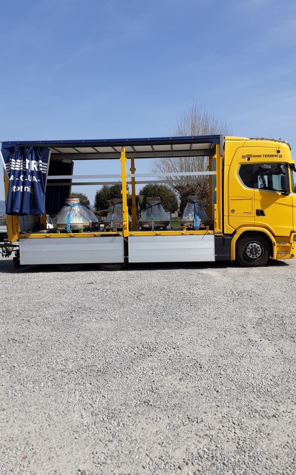 Un camion semirimorchio giallo brillante con telone laterale parcheggiato su una superficie ghiaiosa sotto un cielo azzurro e limpido.
