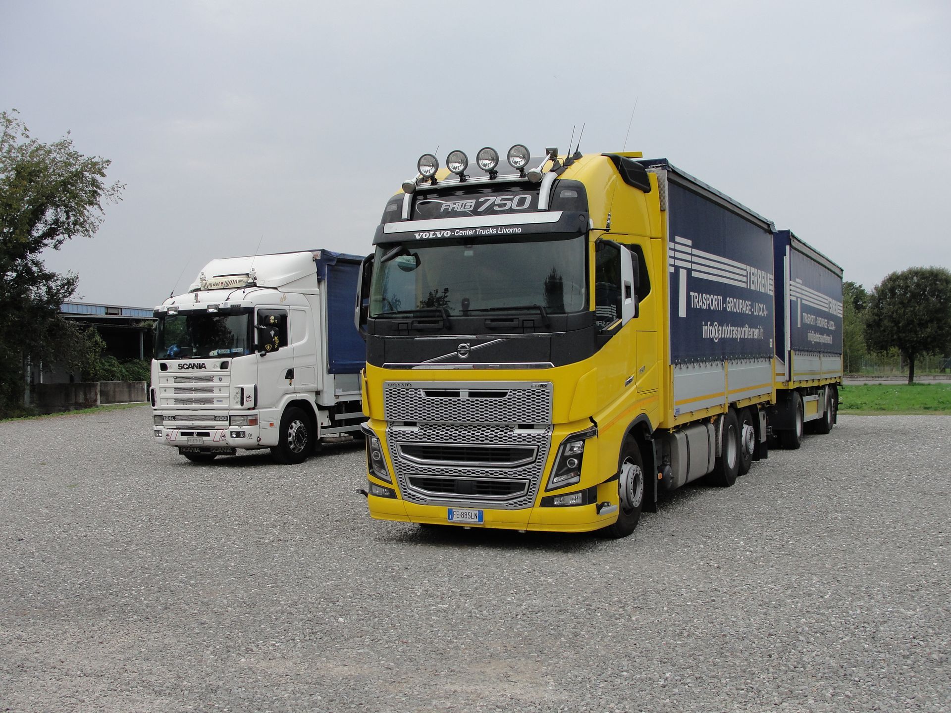 Un camion Volvo giallo e un camion bianco parcheggiati fianco a fianco su un piazzale ghiaioso sotto un cielo nuvoloso.