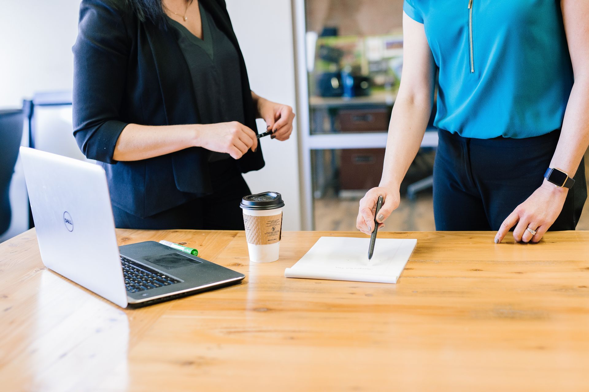 Two women are standing at a table with a laptop and a cup of coffee.