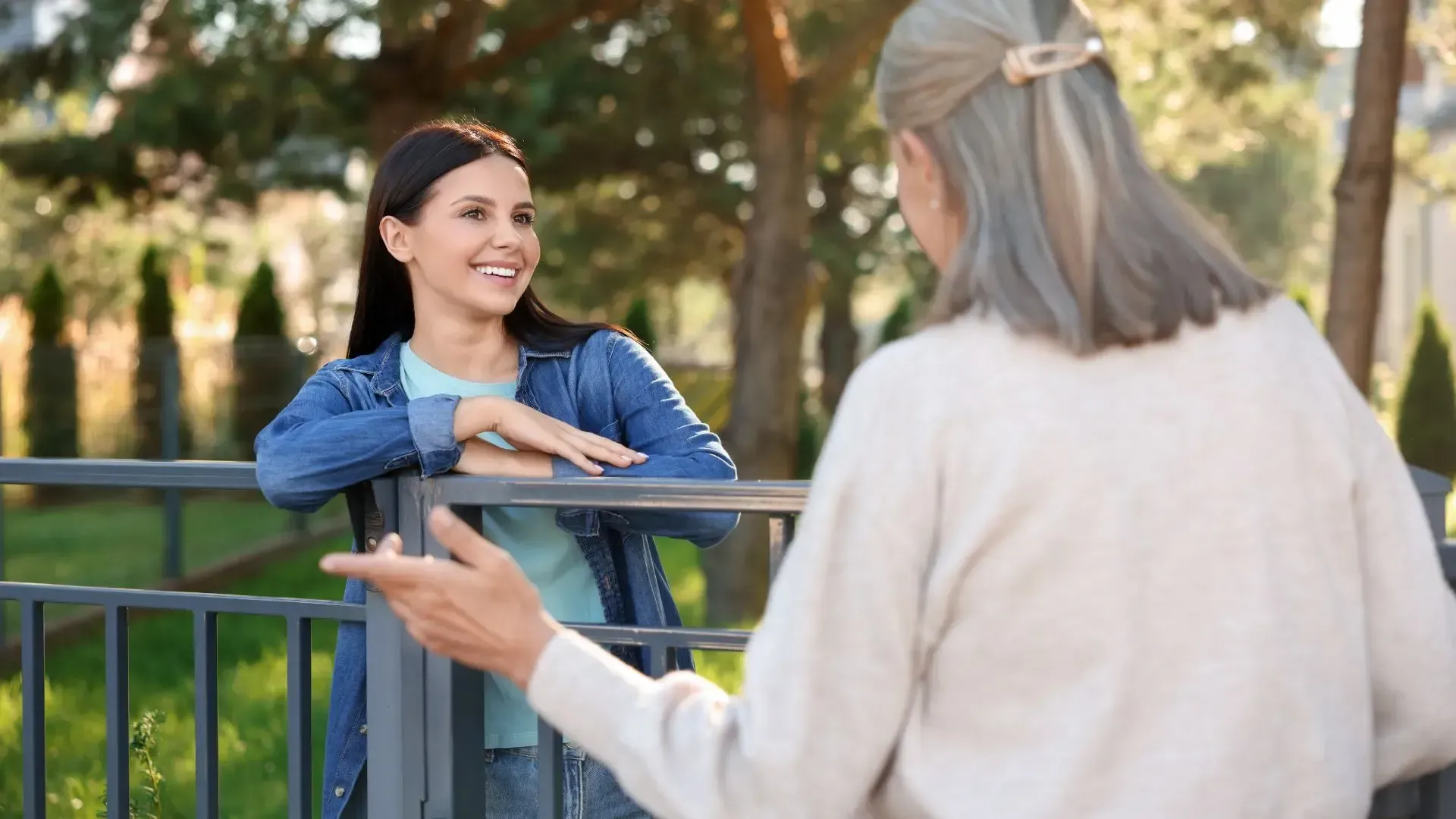 Two women are talking to each other over a fence in a park.