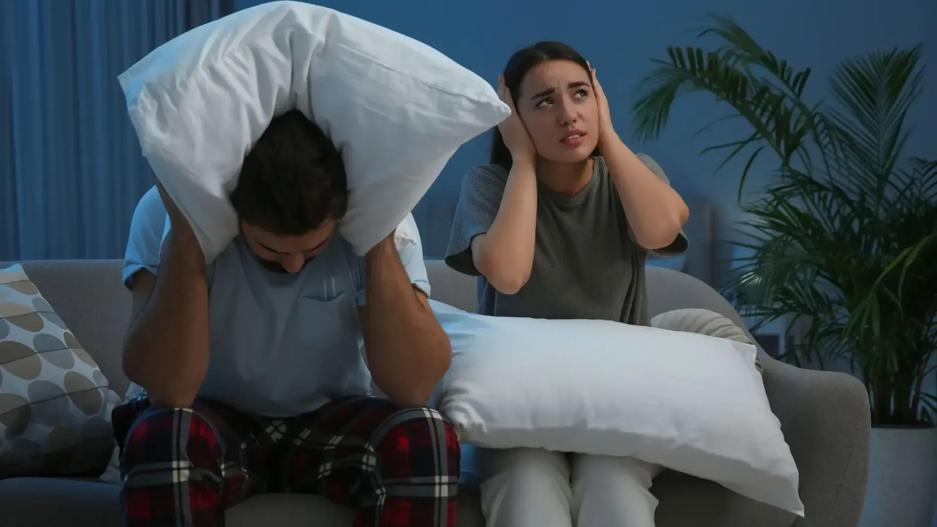 A man and a woman are sitting on a couch covering their ears with pillows.