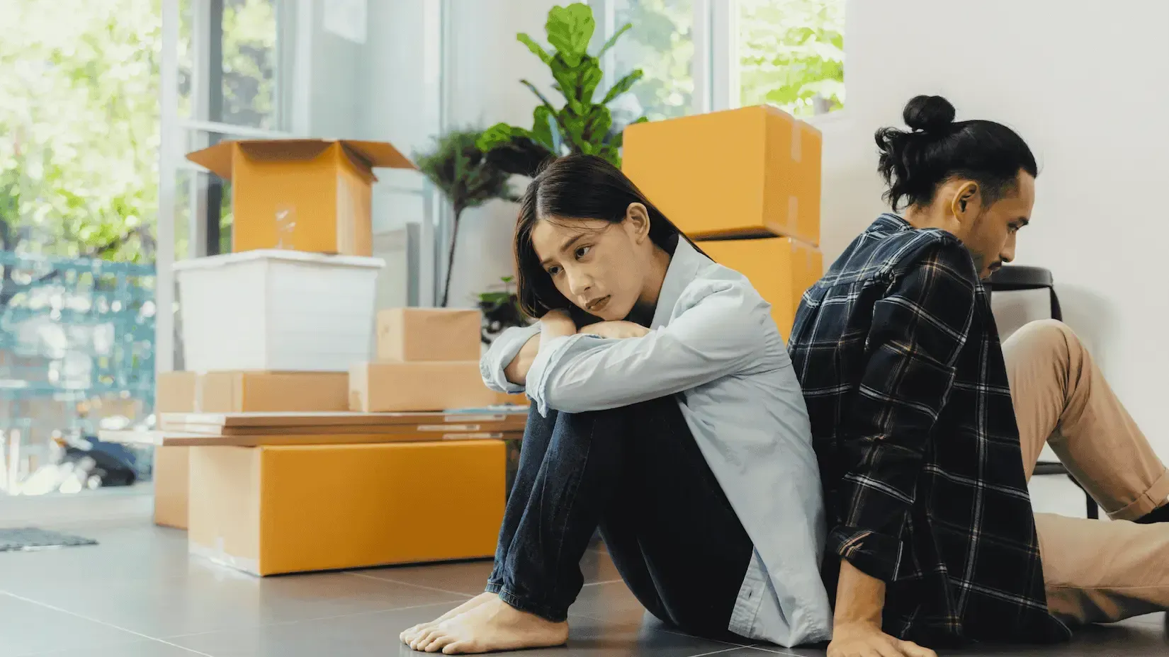 A man and a woman are sitting back to back on the floor in front of moving boxes.