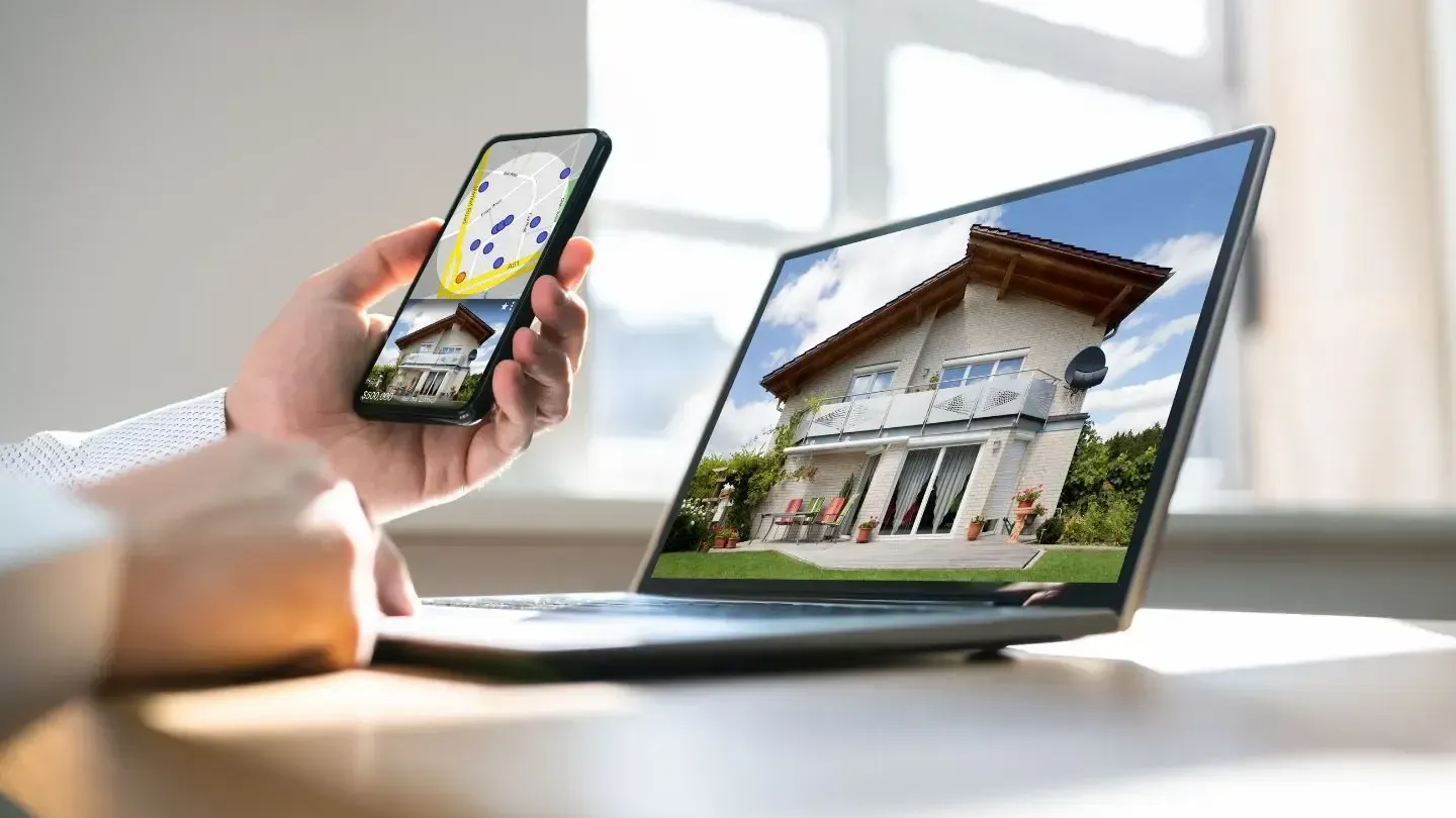 Person uses phone to view a home's details while looking at the same house on a laptop; both screens show the property.
