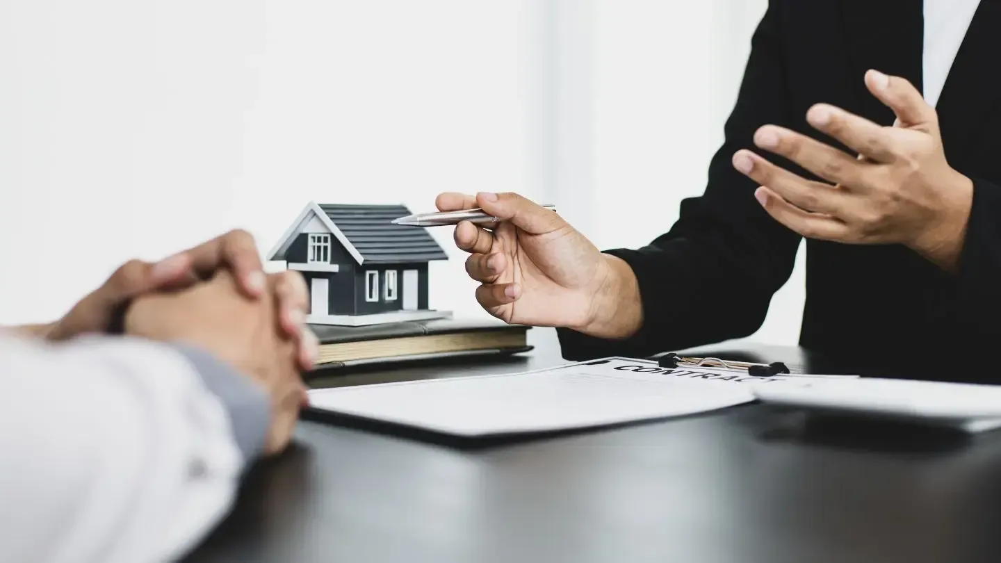 Real estate agent in a black suit gesturing with a pen while talking to a person at a table, with a model house and paperwork visible.