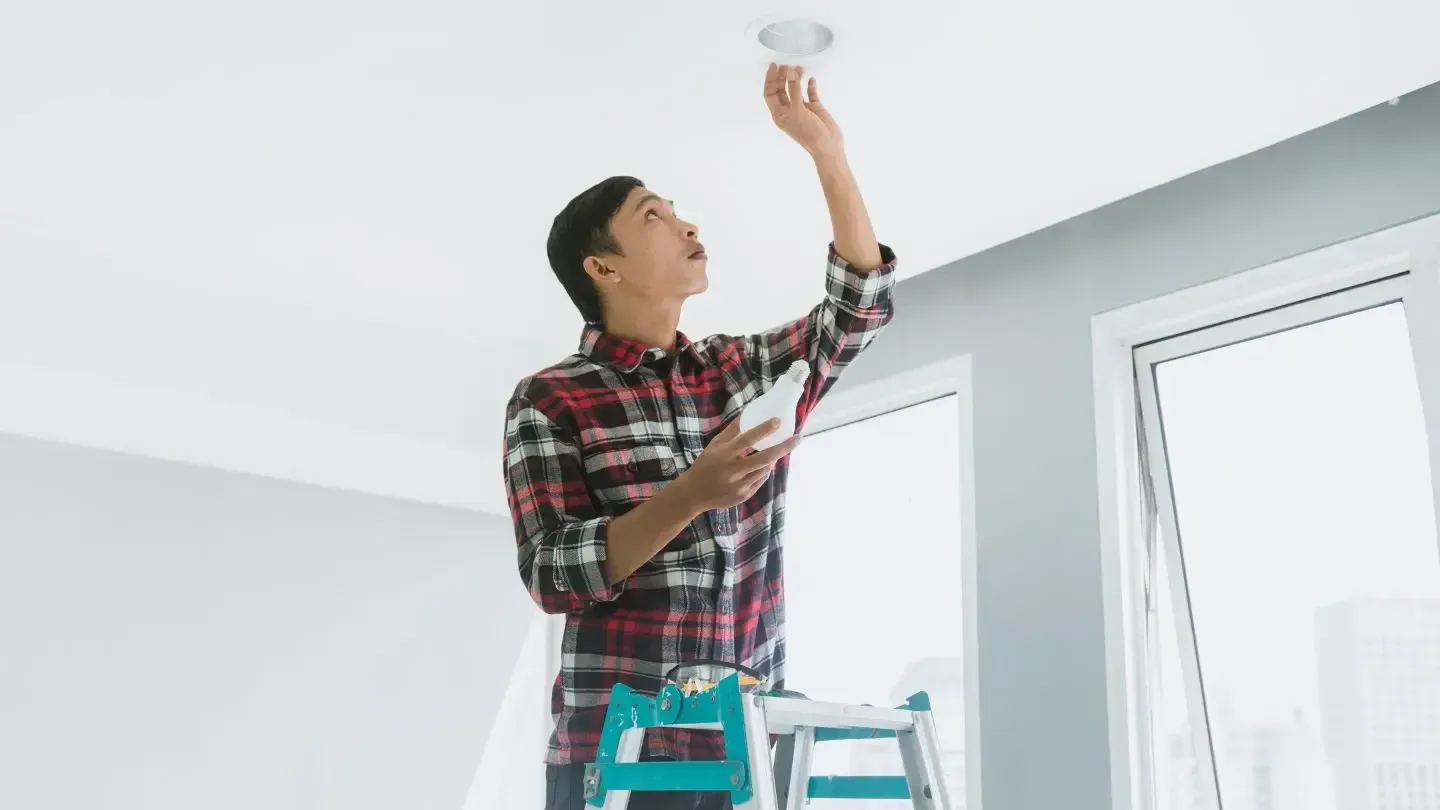 Person on a stepladder installing a recessed light fixture. He is wearing a plaid shirt and looking up.