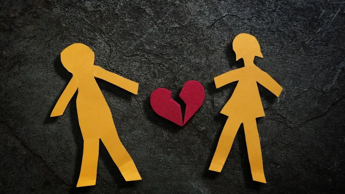 Two paper figures, one male and one female, facing a broken red heart on a black surface.