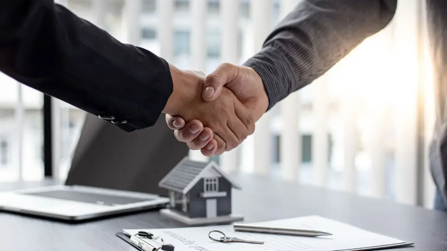 Two people shaking hands over a small house model and contract on a desk.