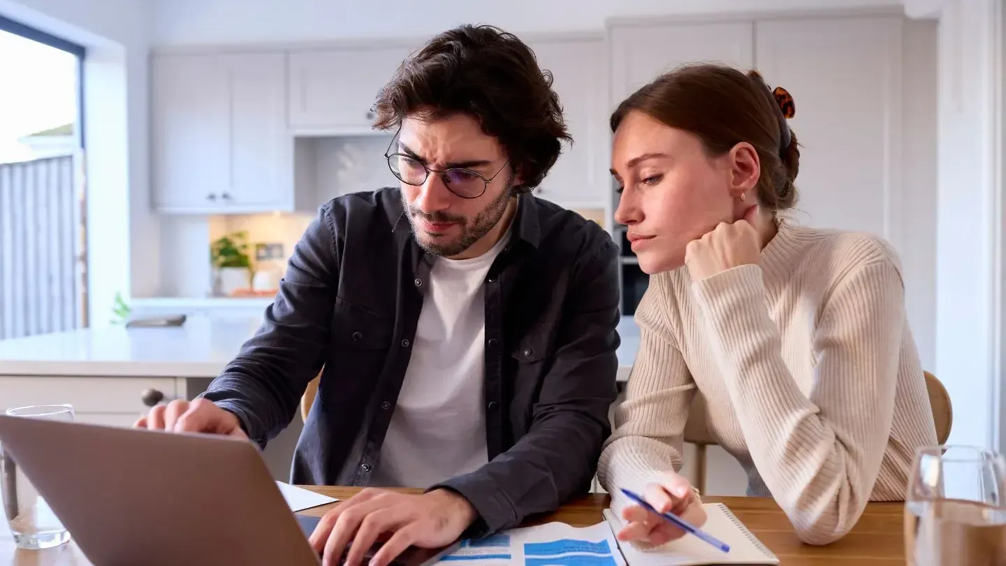 Couple looking at laptop screen together, papers and pens on table. Bright, indoor setting.