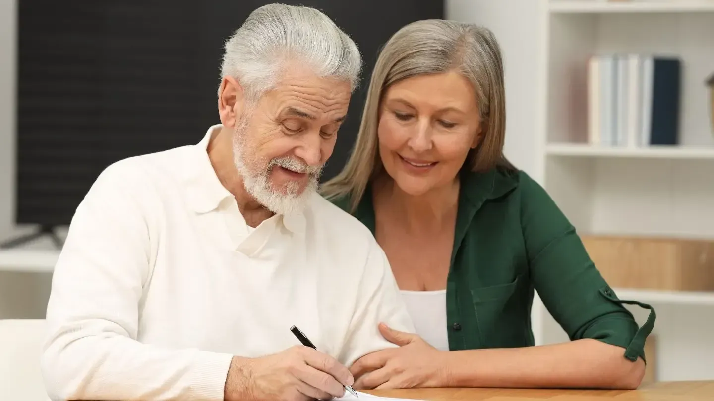 Older person signing paperwork as another person looks on.