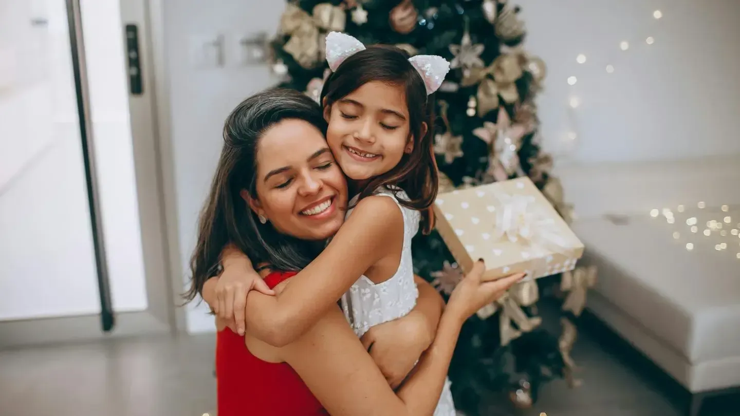 Woman hugging a child, smiling, holding a gift in front of a Christmas tree with gold ornaments and lights.