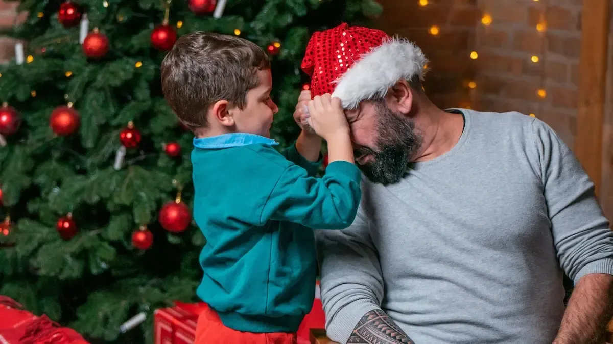 Boy placing a Santa hat on his bearded father in front of a Christmas tree.