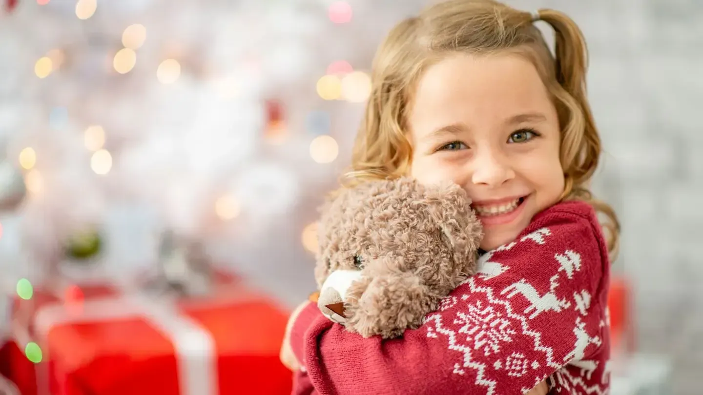 Girl in red sweater hugs teddy bear, smiles in front of Christmas tree and gifts.