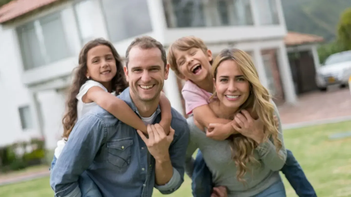 Family of four smiling, piggybacking on parents in front of a house on a sunny day.