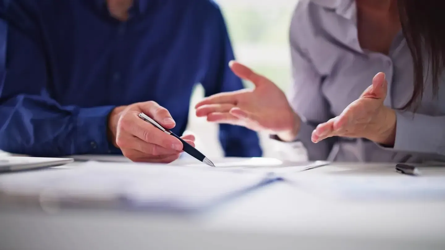 Two people at a table, reviewing documents. One holds a pen and points, the other gestures with hands.