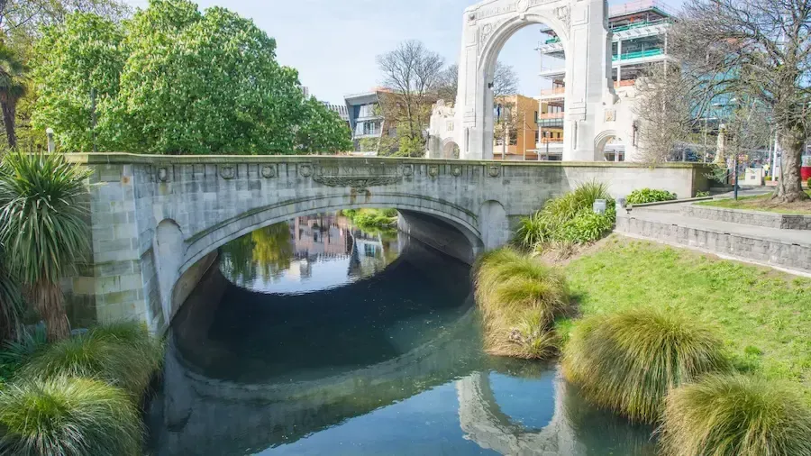 Stone bridge over a calm river in Christchurch, New Zealand. Trees and buildings in the background.
