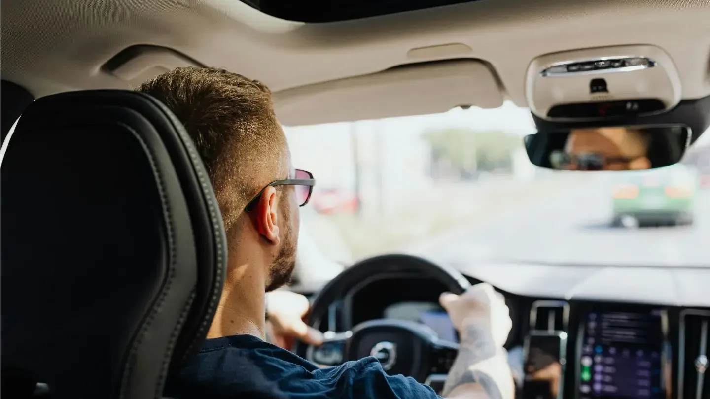 A person in glasses drives a car, seen from the passenger seat, with a view of the road through the front windshield.