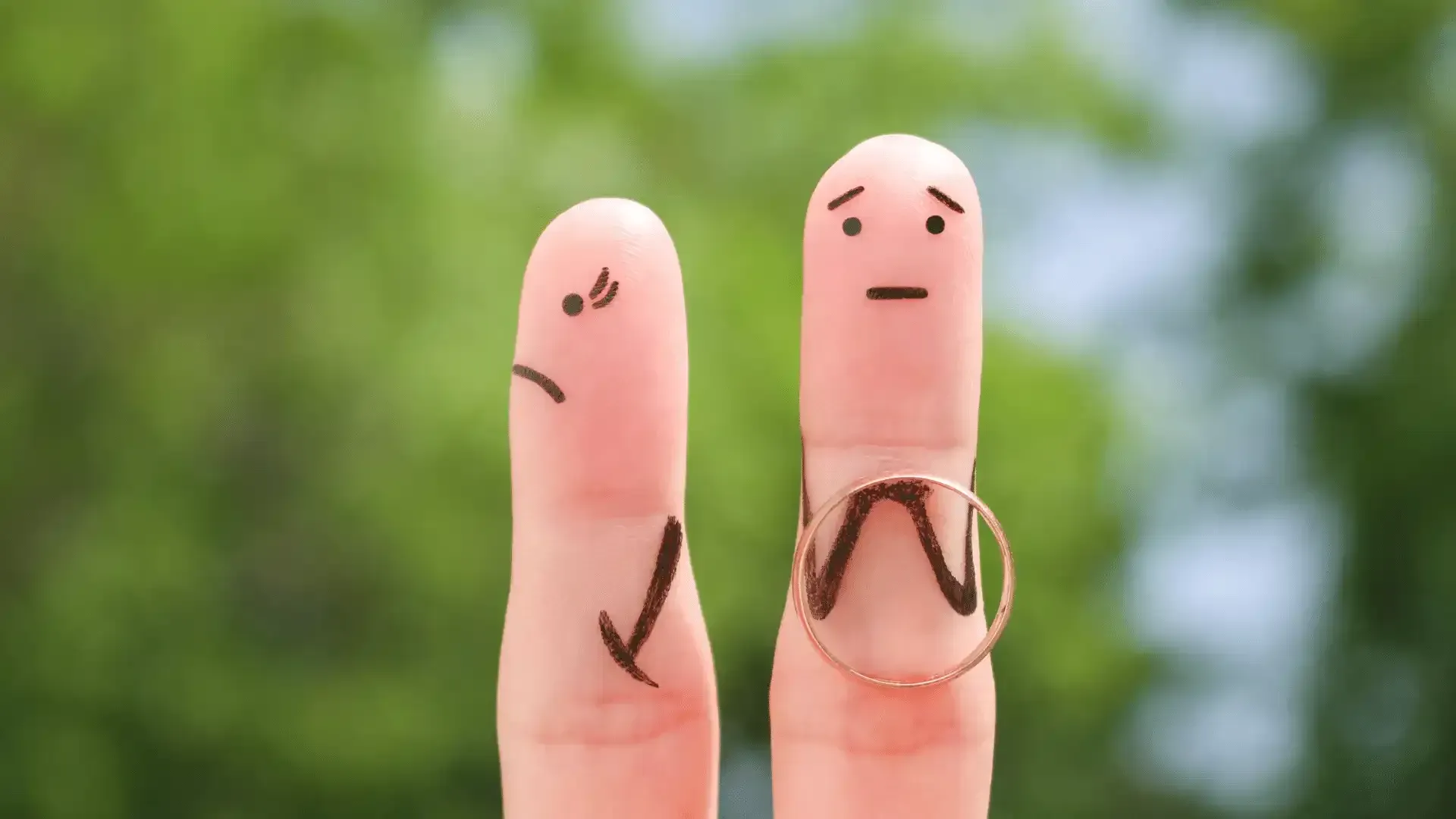 Two fingers with faces drawn on them are holding a wedding ring.