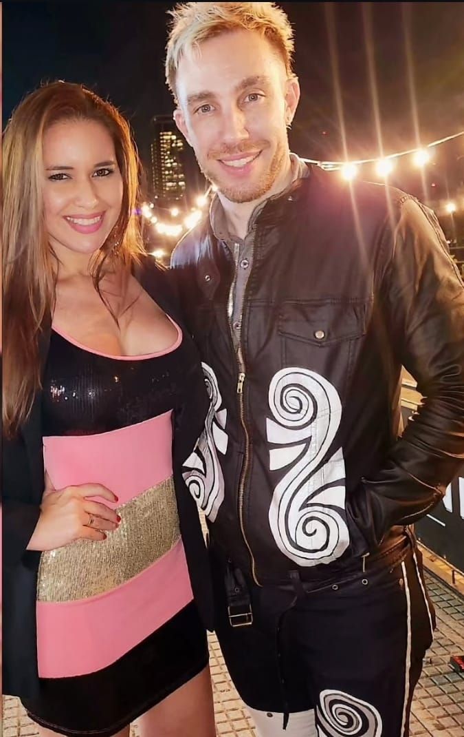 Dos personas posando al aire libre por la noche, vestidas con atuendos negros y rosas, con luces de guirnalda al fondo.