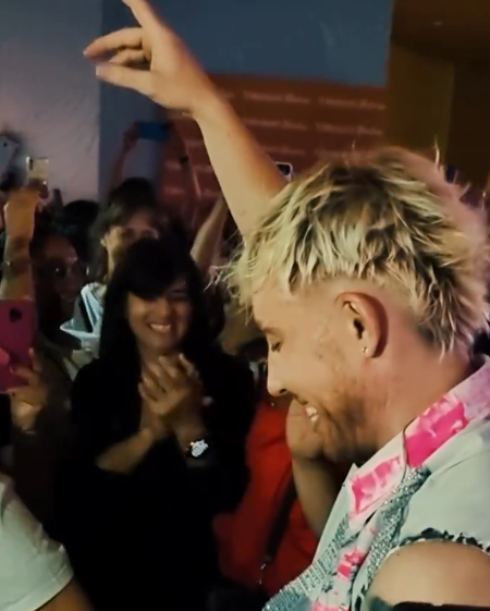 Una persona rubia baila en una fiesta abarrotada, sonriendo con el brazo levantado; se ve una camisa rosa con estampado floral.