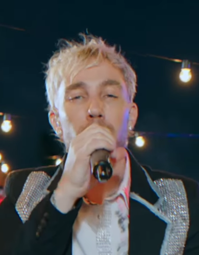 Cantante rubia sosteniendo un micrófono en un escenario con luces colgantes, vistiendo una chaqueta brillante.