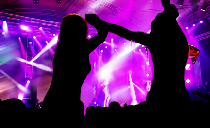 Dos aficionados, silueteados, alzan las manos en un escenario de concierto iluminado de color púrpura con potentes focos.