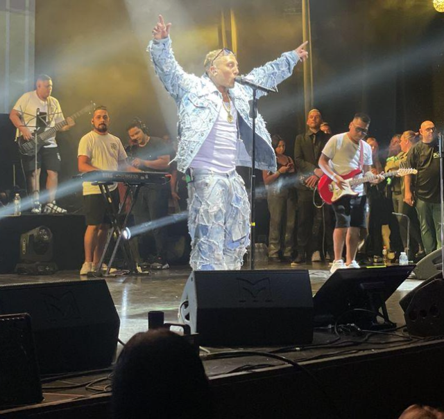 Un cantante con un traje azul levanta los brazos en un escenario de concierto con una banda y luces brillantes detrás.