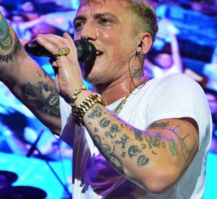 Cantante con brazos tatuados actuando frente a un micrófono en el escenario bajo luces azul-violeta.