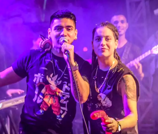 Dos artistas en el escenario bajo una iluminación púrpura; uno canta en un micrófono mientras el otro sostiene una maraca roja.