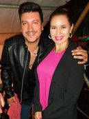 Dos personas sonrientes posando juntas en interiores, una con una chaqueta de cuero negra y la otra con un blazer negro y una blusa rosa.