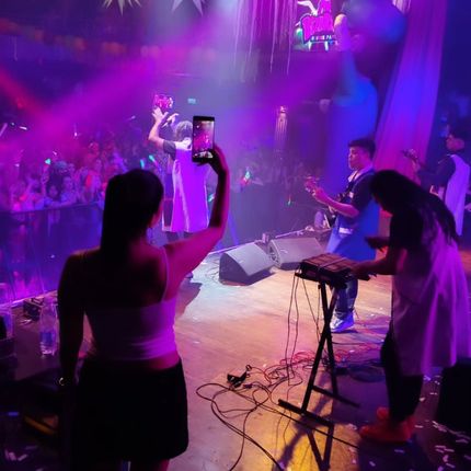 Una cantante actuando en un escenario iluminado con luces moradas, con un público filmando y un teclista cerca.