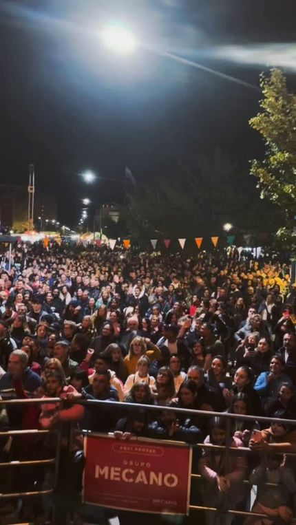 Una multitud se reunió en un evento nocturno al aire libre bajo luces brillantes, con pancartas rojas de MECANO en la entrada.