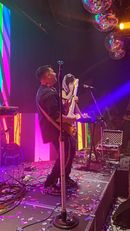 Músicos actuando en un escenario colorido con confeti e iluminación púrpura.