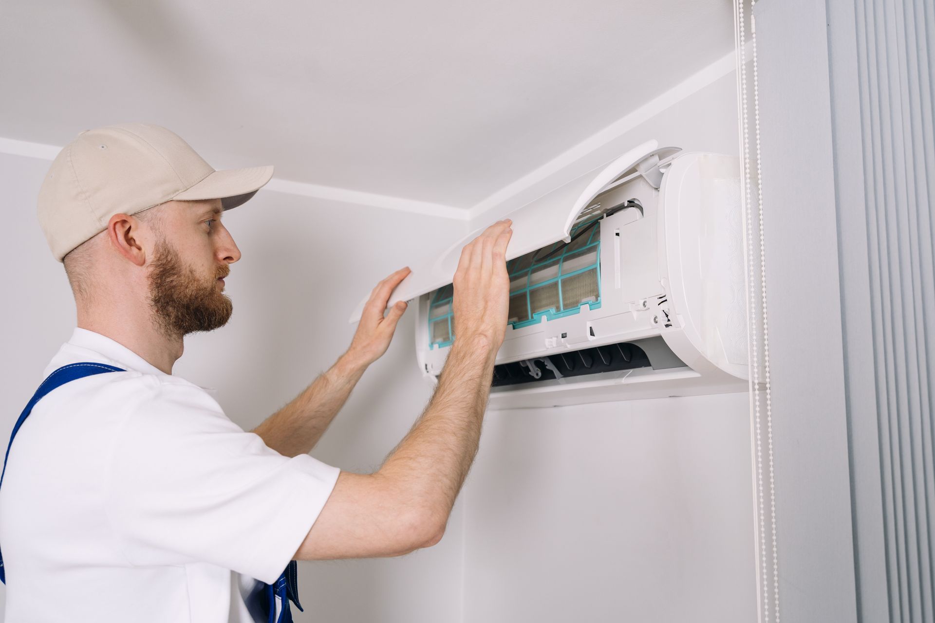 Technician in white uniform and cap removing the filter panel of a wall-mounted mini-split AC unit.