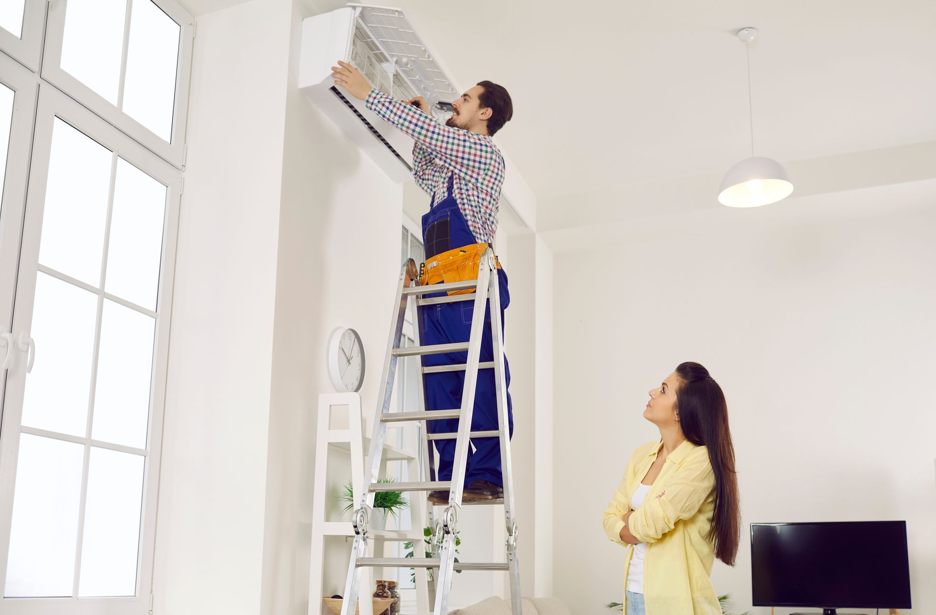 A technician on a ladder is servicing a wall-mounted AC unit indoors.