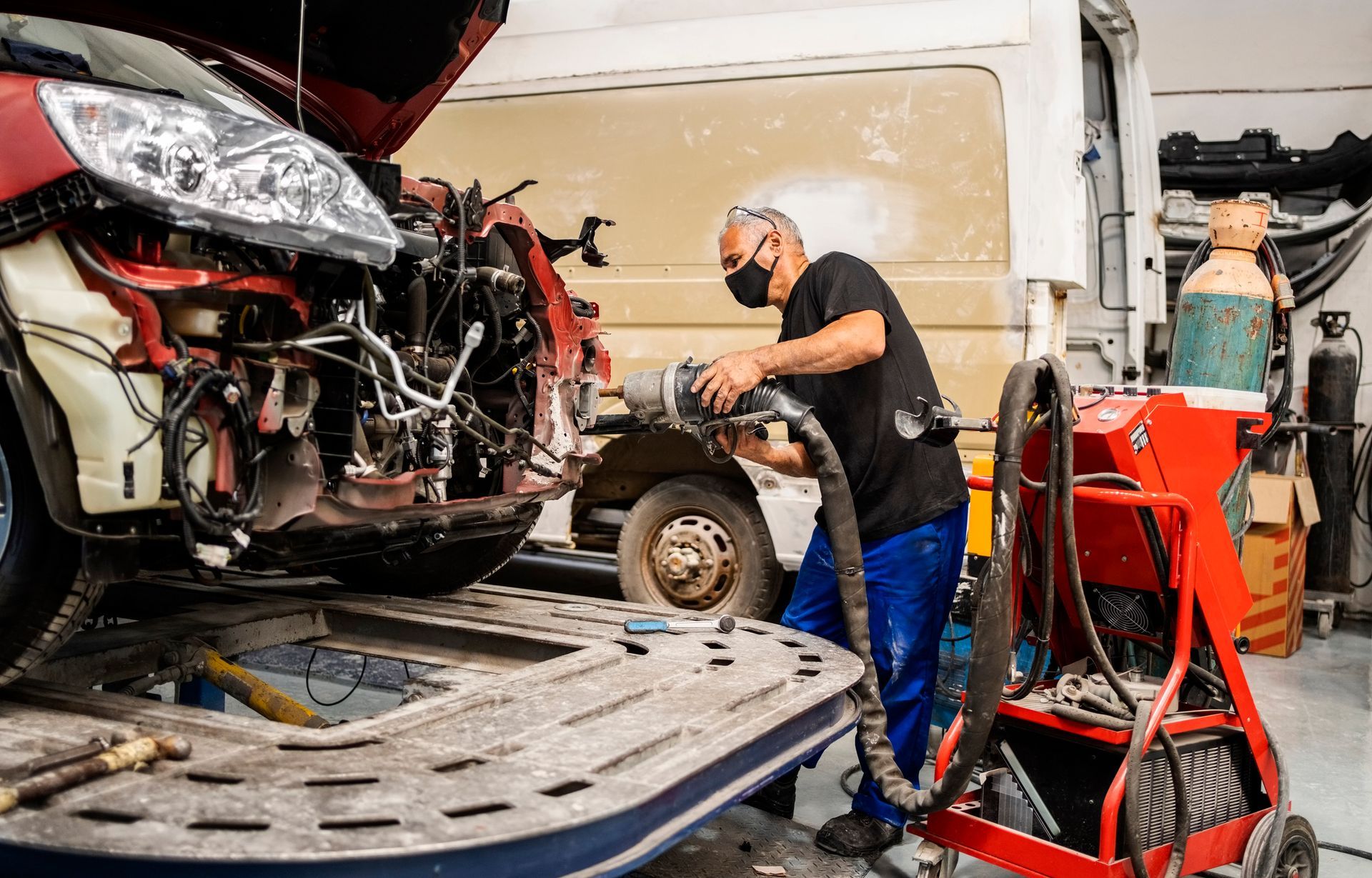 Mechanic in mask repairs a damaged red car in a garage.