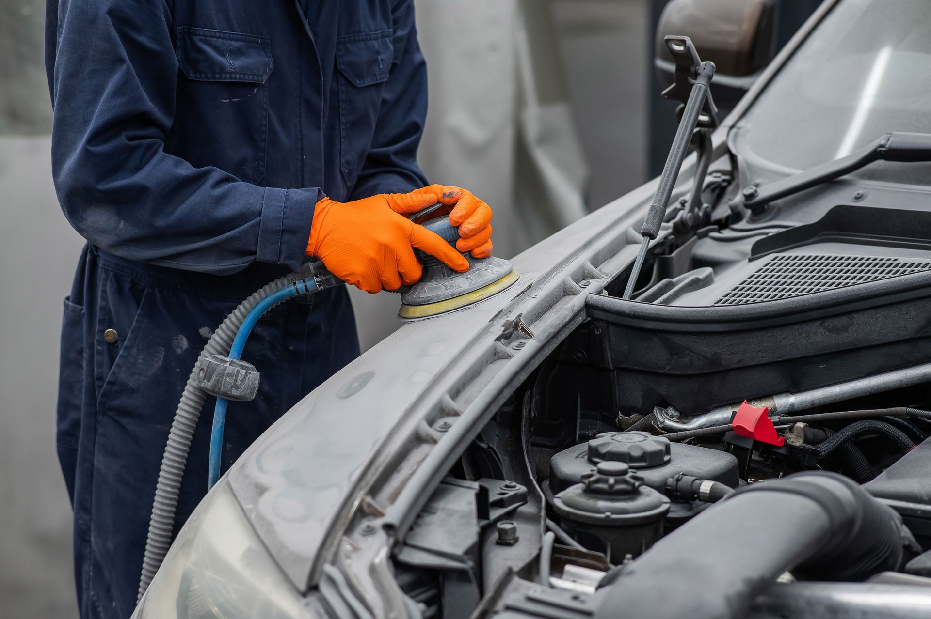 Person in blue coveralls sands a car hood with an orange-gloved hand, engine bay open.