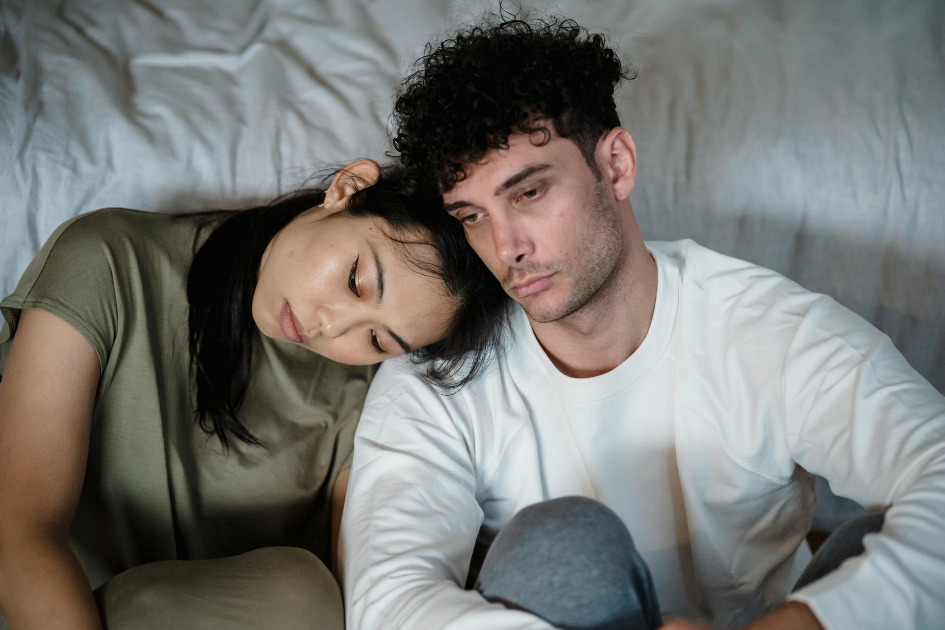 Woman resting head on man's shoulder; both appear sad, seated on a bed.