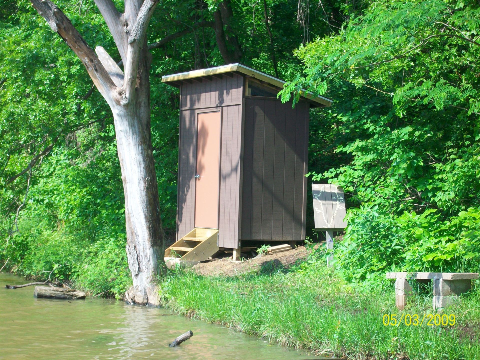 A small shed is sitting next to a tree near a body of water.