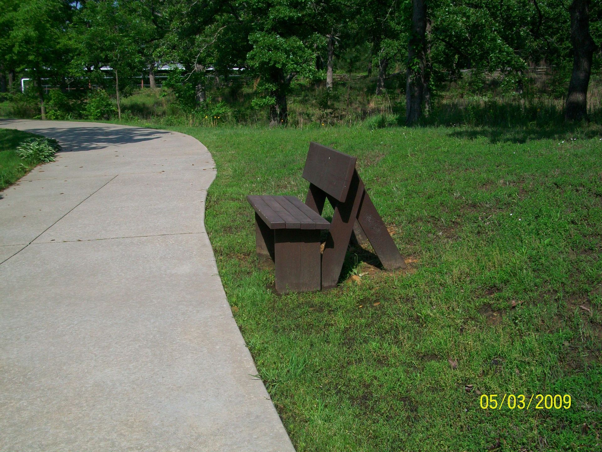 A wooden bench sits in the grass next to a sidewalk