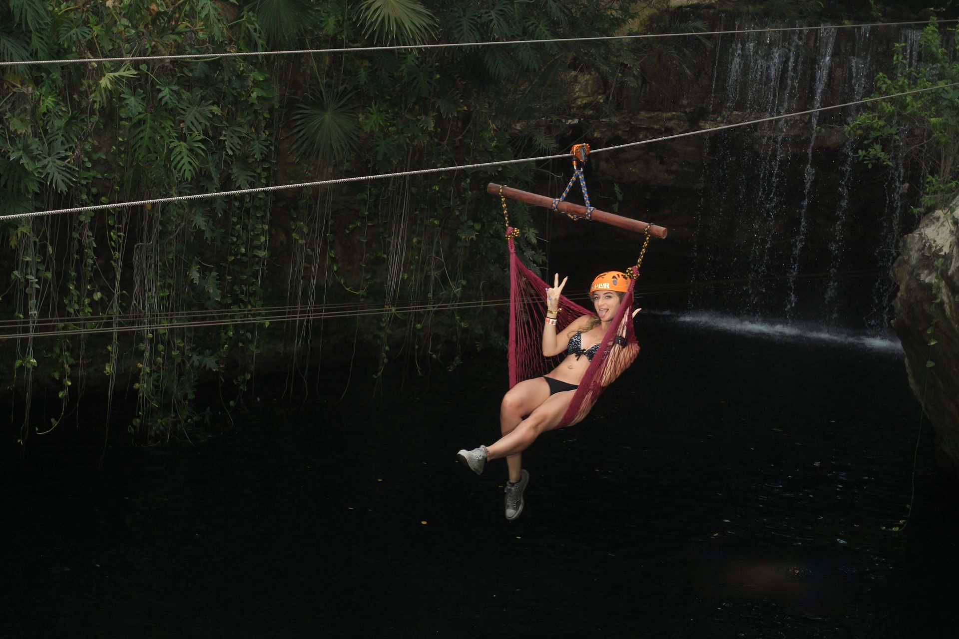 Woman in a swimsuit on a swing over a body of water, near a waterfall, with peace signs.